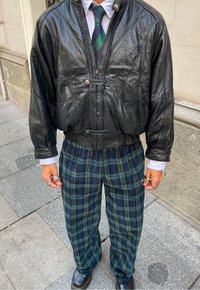 Chaqueta bomber de cuero negro con una cintura ajustada, combinada con pantalones de cuadros verdes y marino de pierna ancha y zapatos de plataforma negros.