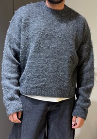 Homme portant un pull gris duveteux par-dessus une chemise blanche et un jean sombre ample, debout à l'intérieur devant un fond uni.