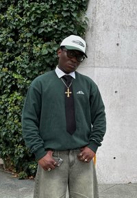 Green sweatshirt with a logo, white collared shirt, black tie, and light grey wide-leg jeans. Accessories include a cross necklace and rings.