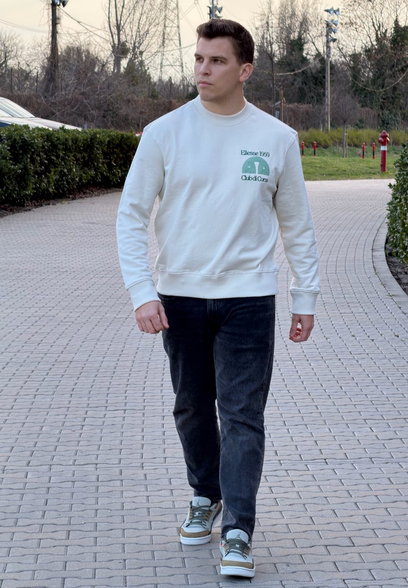 Jauns vīrietis staigā pa bruģētu taku baltā džemperī, melnās biksēs un sporta apavos, apjozts ar krūmiem un kokiem vēlā pēcpusdienā.