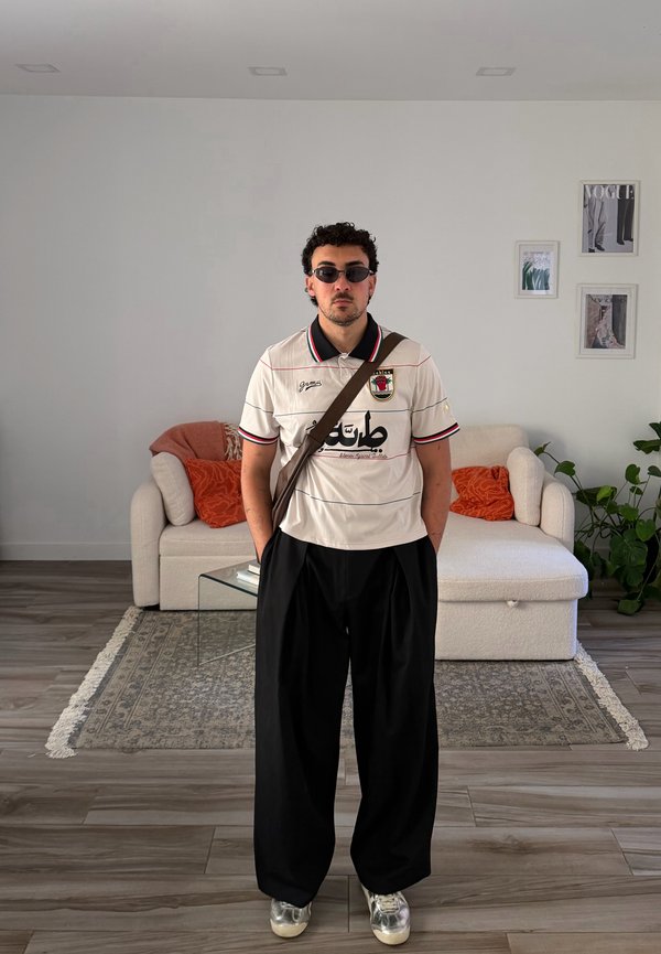 Ein Mann mit Sonnenbrille, einem weißen Poloshirt mit arabischem Schriftzug und schwarzen weiten Hosen steht in einem Wohnzimmer mit einem beigen Sofa und Pflanzen hinter sich.