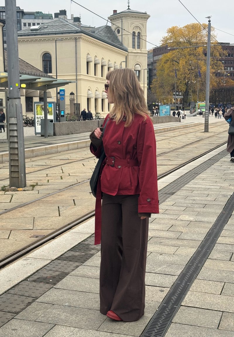 Red belted jacket over a dark shirt, paired with brown pinstripe wide-leg pants and red shoes, set on a city street with tram tracks.