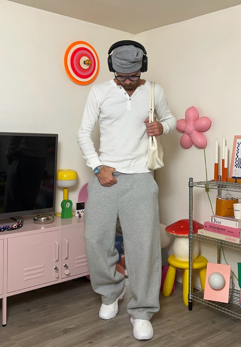 Maglietta henley bianca a maniche lunghe, pantaloni della tuta grigi, sneakers bianche, berretto grigio e cuffie nere. Borsa beige e decorazioni di sfondo colorate.