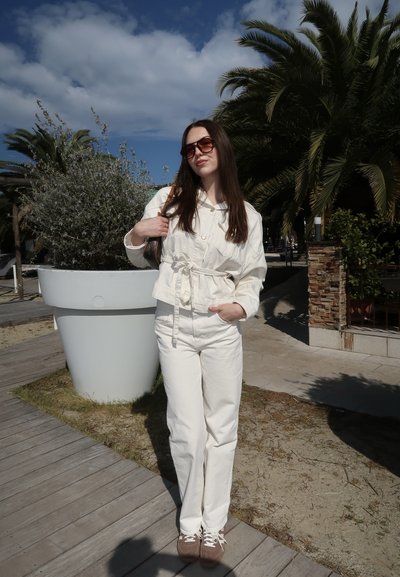 Mujer con ropa blanca y gafas de sol de pie sobre un camino de madera junto a una gran maceta blanca con plantas de hojas y palmeras bajo un cielo azul.