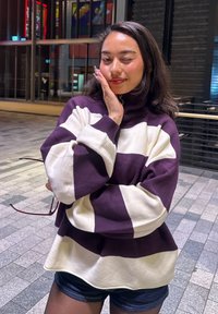 Ylisuuri neulepaita violeteissa ja kermaisissa raidoissa, jossa on korkea kaulus ja resorit hihansuissa. Malli pitelee aurinkolaseja, ja neuleen kangas on kuvioitua.