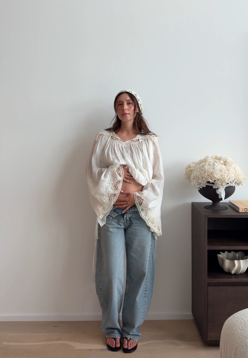 Femme en blouse blanche et foulard se tient près d'une étagère, les mains sur le ventre, à côté d'un vase de fleurs blanches et d'un livre dans une pièce minimaliste.