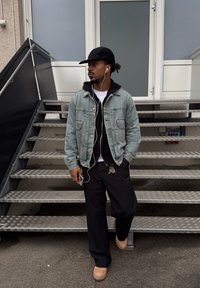 Homme portant une casquette noire, une veste en denim et des écouteurs, debout sur des escaliers tenant un téléphone, regardant sur le côté à l'entrée d'un bâtiment.