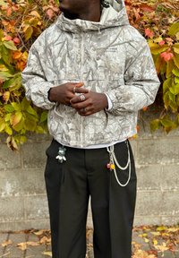 Svetlá bunda na zips s maskáčovým vzorom, s kapucňou a elastickými manžetami. Čierne nohavice s príveskami na kľúče a prstene na rukách.