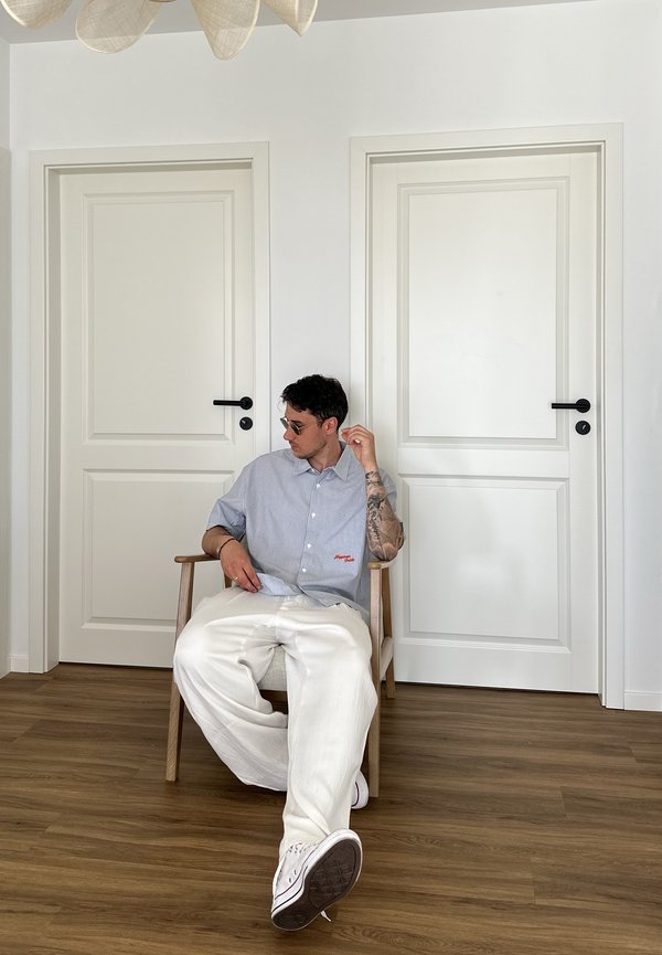 Man met zonnebril en casual kleding zit op een houten stoel tussen twee gesloten witte deuren op een houten vloer.