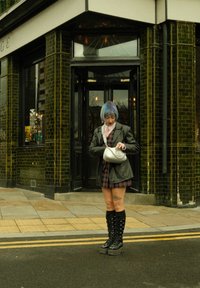 Ένα άτομο στέκεται έξω από ένα κτίριο, φορώντας ένα μαύρο δερμάτινο μπουφάν, καρό φούστα, μαύρες μπότες μέχρι το μηρό και κρατώντας μια ασημένια τσάντα.