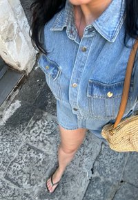 Σετ από τζιν πουκάμισο και σορτσάκι σε ανοιχτό μπλε χρώμα, με χρυσές φερμουάρ και δύο τσέπες στο στήθος. Υφασμάτινη καφέ τσάντα; σανδάλια στα πόδια. Πέτρινη πλάκα από κάτω.