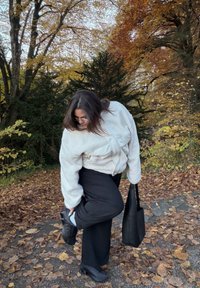 Jachetă supradimensionată din fleece alb cu material texturat, pantaloni negri cu picioare largi și adidași negri. Geantă de umăr întunecată în mână; frunze de toamnă în fundal.