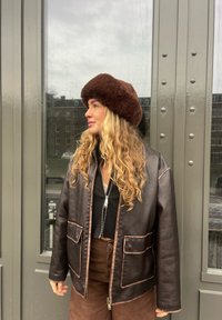 Giacca di pelle marrone con cuciture chiare a contrasto e due tasche frontali, abbinata a un cappello di pelliccia marrone. Sullo sfondo si vedono finestre grigie.