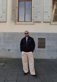Schwarze Zip-Jacke, gemustertes blaues Hemd, beigere weit geschnittene Hose und schwarze Schuhe. Steht vor einer Steinmauer und einem Fenster.