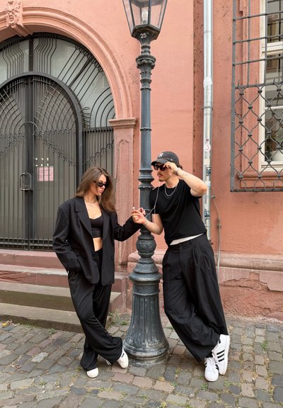 Dos personas con ropa negra y gafas de sol comparten auriculares mientras se apoyan en una farola en una calle empedrada.