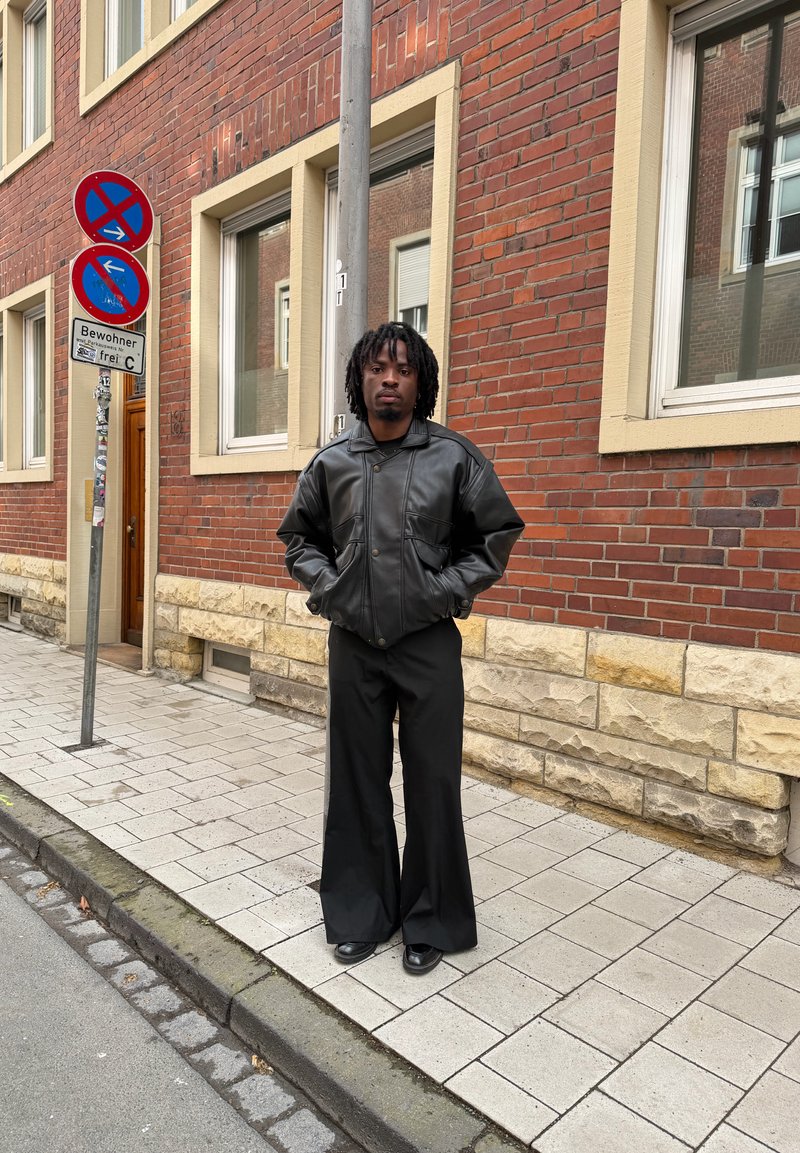 Homme portant une veste en cuir noire et un pantalon noir large, debout sur un trottoir à côté d'un bâtiment en briques et de panneaux de signalisation.