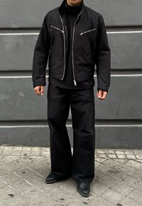 Uomo vestito con una giacca nera con zip e tasche sul petto, una camicia nera, pantaloni larghi neri e scarpe nere, in piedi su un marciapiede piastrellato.