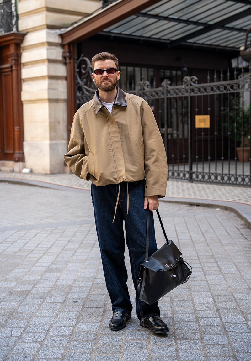 Homme portant une veste beige, un jean foncé et des lunettes de soleil, se tient dans une rue pavée en tenant un grand sac en cuir noir.