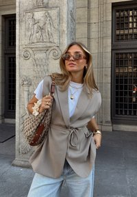 Bežna tkaninska jakna s trakom za pas, složena preko bele majice. Dodatci uključuju naočare za sunce, slojevite ogrlice i brašnasti torbu sa uzorkom.