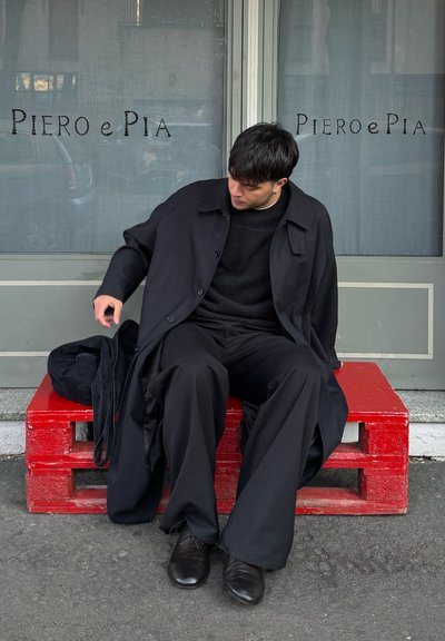 Joven con ropa negra sentado en un banco hecho de palets rojos frente a una tienda con el letrero "Piero e Pia" en la ventana detrás de él.