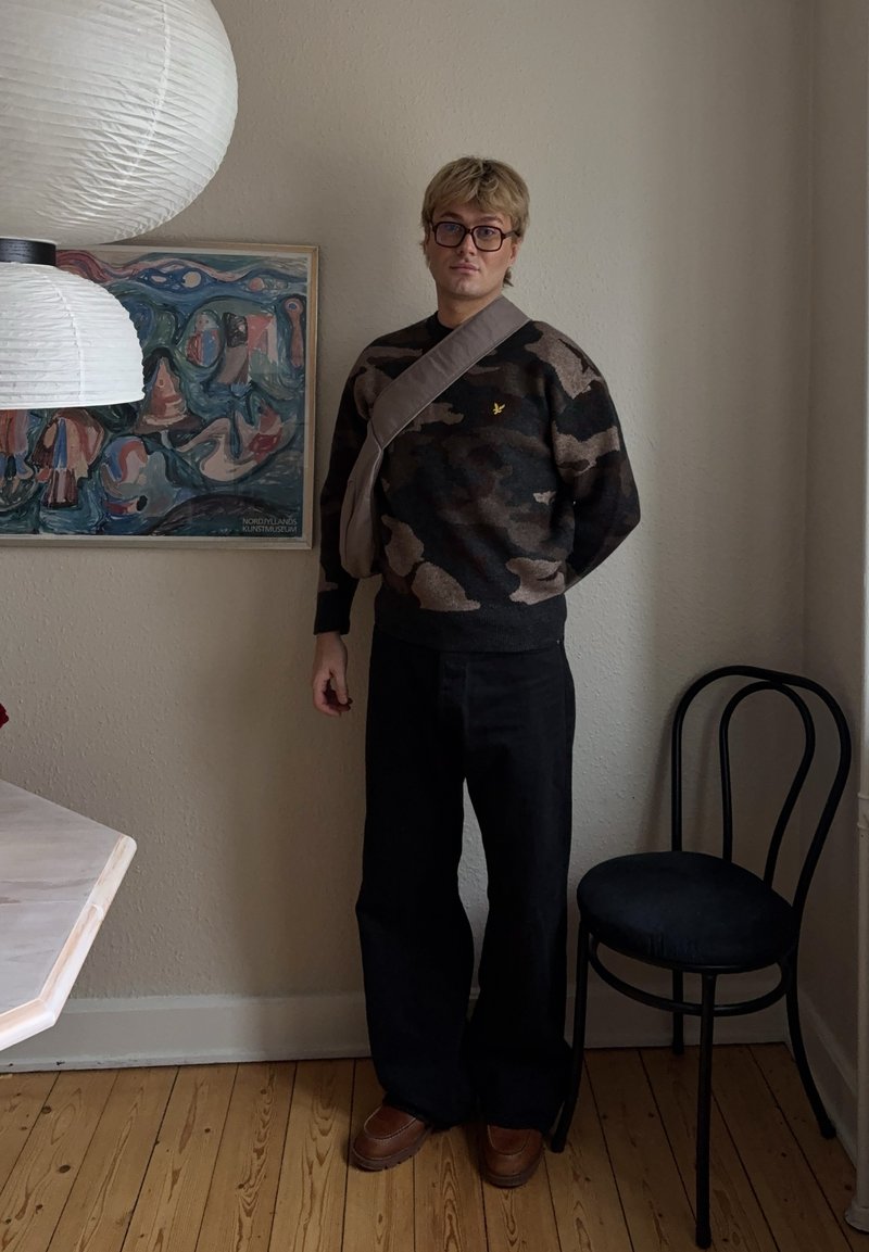 Personne aux cheveux blonds courts et aux lunettes, debout à l'intérieur, portant un pull camouflage, un pantalon noir, des chaussures marron et un sac en bandoulière.