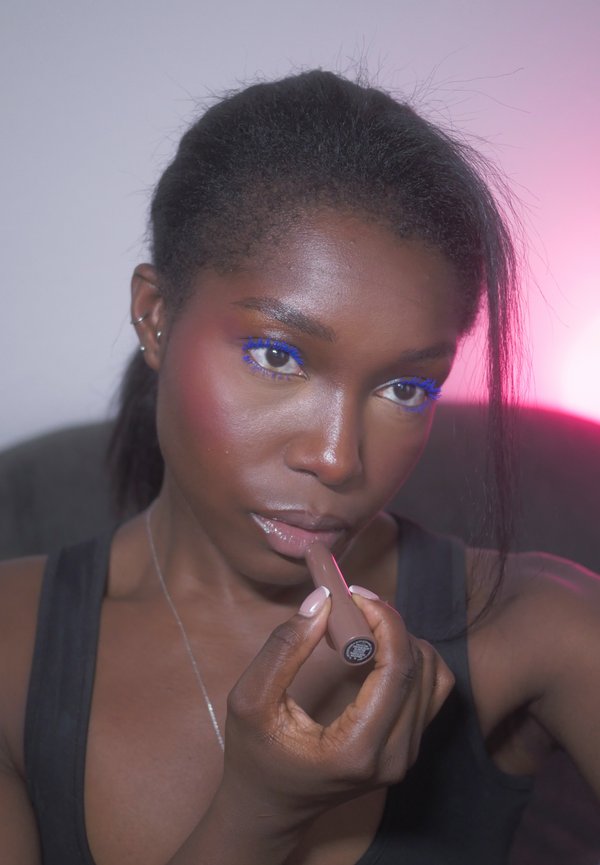 Une femme applique un gloss rose et porte du mascara bleu vif, avec un éclairage rose de studio derrière elle.