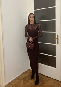 Femme en longue robe noire en dentelle tenant un petit sac à main marron, debout à l'intérieur près d'une porte blanche avec des panneaux de verre dépoli.