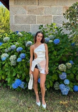 Vestido blanco de un solo hombro con cortes con acentos texturizados, combinado con pantalones cortos negros y zapatillas blancas. Bolsa de mimbre. Rodeada de hortensias azules.