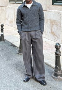 Pull-over en tricot gris foncé avec col, assorti à un pantalon large gris rayé et des chaussures noires, sur un fond en pierre.