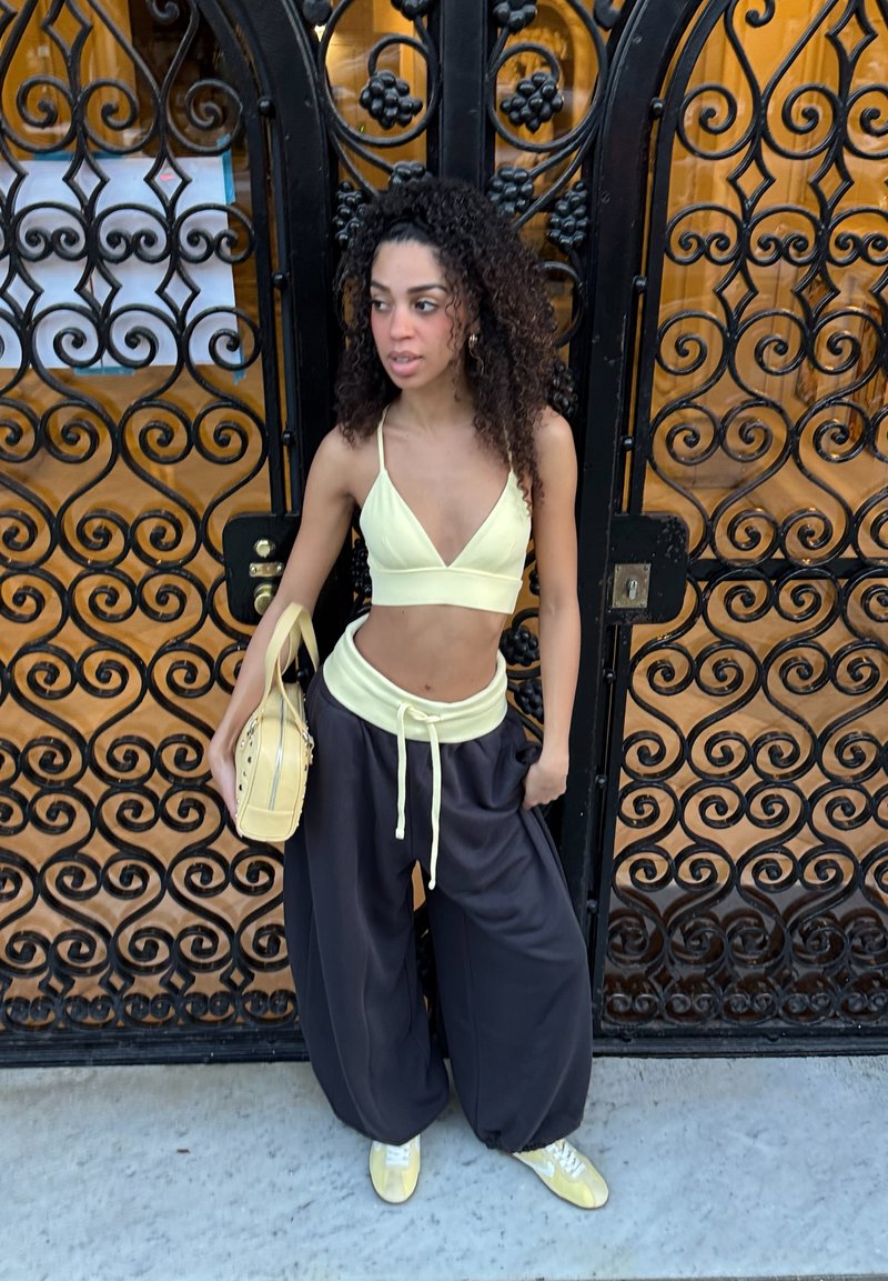 Jeune femme aux cheveux bouclés portant un crop top jaune, un pantalon noir ample, des baskets jaunes, et tenant un sac à main jaune, debout près d'une porte noire ornée.