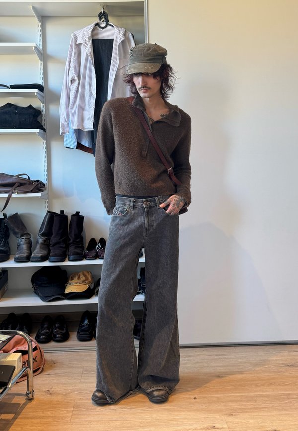 Junge Person in braunem Pullover, grauen weiten Jeans und Mütze steht in einem Raum mit Regalen, in denen Stiefel, Schuhe und gefaltete Kleidung stehen.