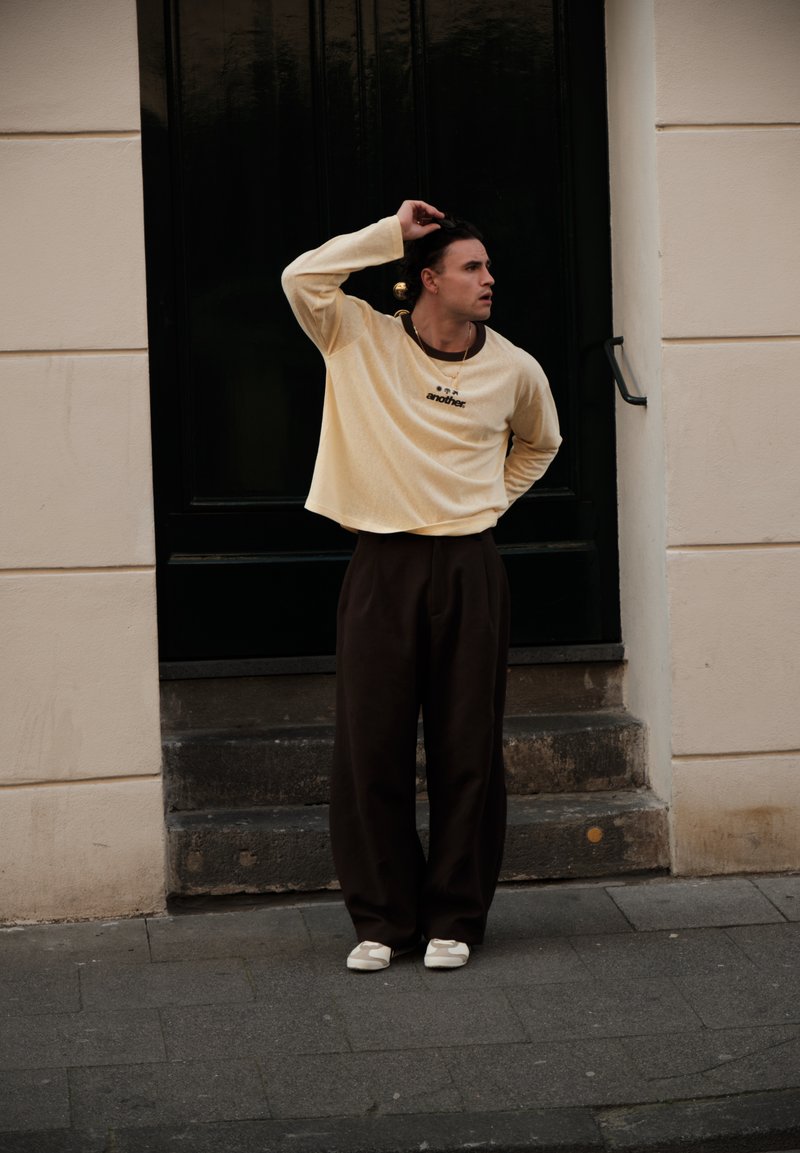 Homme aux cheveux foncés, portant une chemise beige à manches longues et un pantalon large foncé, debout sur le trottoir, se touchant la tête, devant une porte noire et des marches en pierre.