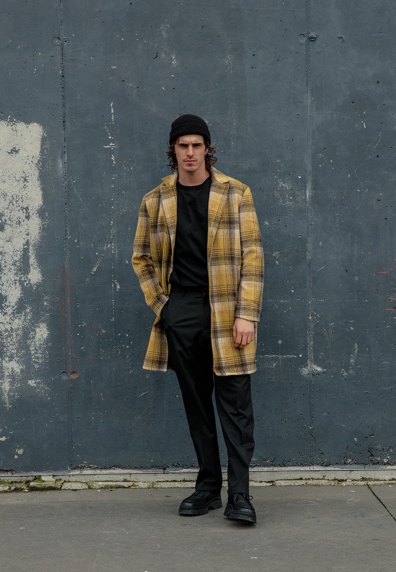 Geltonas čekeruotas paltas su juodais akcentais, dėvimas virš juodos marškinėlių, derinamas su juodomis kelnėmis ir masyvais batais; tekstūruotas audinys.