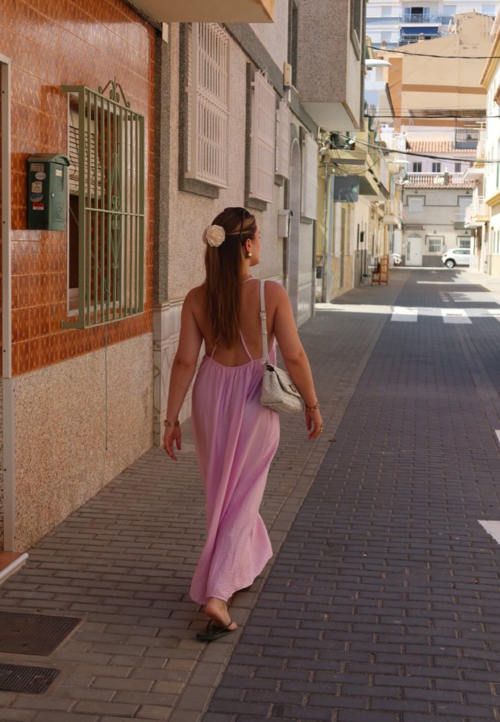 Ροζ, αμάνικο maxi φόρεμα με ανοιχτή πλάτη, περπατώντας σε μια πλακοστρωμένη οδό; φοράει ένα κρεμ αξεσουάρ μαλλιών και κρατάει μια τσάντα.