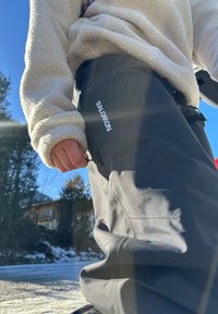Pantalon de ski noir avec une texture lisse et une poche zippée latérale, arborant un logo "Salomon" blanc. Un pull en polaire crème est visible en haut.