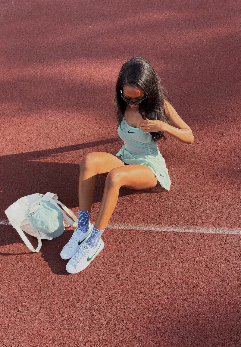 Jeune femme en tenue de sport assise sur la piste rouge, ajustant son haut, avec un sac blanc et une casquette à côté d'elle.