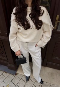 Jasno beżowy, oversize sweter, białe, plisowane spodnie oraz mała czarna torebka. Ciemne, spiczaste buty i fakturowane, brązowe tło.