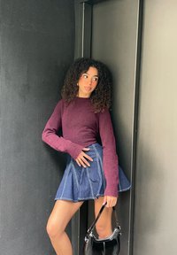 Femeie cu păr creț purtând un pulover mov și o fustă albastră, ținând o geantă neagră, stând în fața unui perete texturat gri.