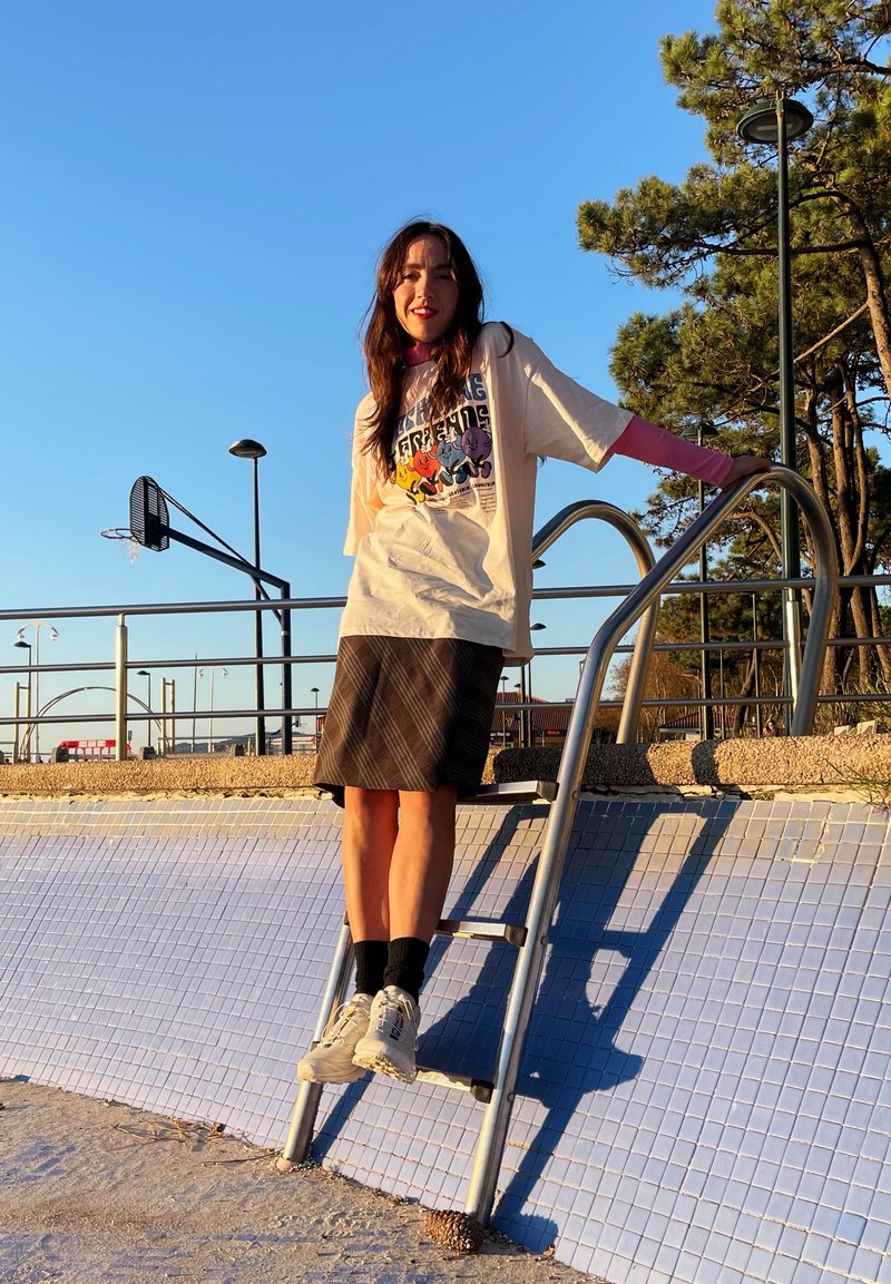 Μεγαλόσωμο κρεμ γραφικό t-shirt με πολύχρωμους χαρακτήρες, συνδυασμένο με μια μαύρη φούστα με σχέδια. Λευκά αθλητικά παπούτσια και μαύρες κάλτσες ολοκληρώνουν την εμφάνιση.
