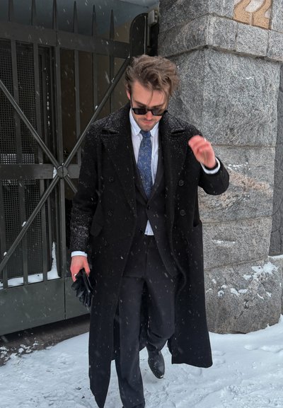 Hombre con abrigo negro, traje y corbata caminando bajo la nieve cerca de un muro de piedra y una puerta de metal, sosteniendo guantes negros en una mano.