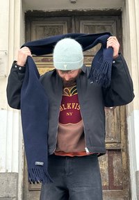 Námořnický vlněný šál s třásněmi, který je drženo vysoko. Osoba má na sobě černou bombera nad hnědým svetr s vínovým a zlatým nápisem.