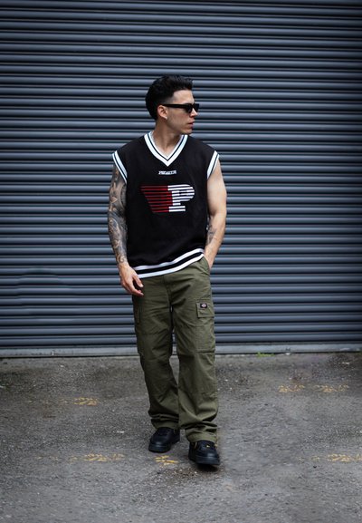 Jersey sin mangas negro con logo de rayas blancas y rojas, combinado con pantalones cargo verde oliva y zapatillas negras, contra un fondo gris.