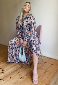 Floral maxi dress featuring a blend of blues, oranges, and pinks. Complemented by a light blue handbag and lavender sandals. Set against a wooden floor background.