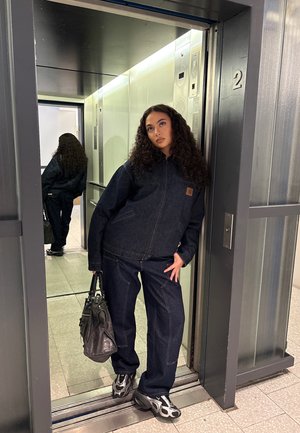 Dark denim jacket and pants, featuring a patch logo. Black bag in hand; wearing gray and black sneakers. Elevator setting with mirror.