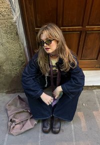 Τζάκετ από τζιν και παντελόνι σε φαρδιά γραμμή σε σκούρο μπλε, oversized σχέδιο. Καφέ δερμάτινα μποτίνια μέχρι τον αστράγαλο, με ένα ανοιχτόχρωμο πουκάμισο και μια μοβ γραβάτα. Χαλαρή καφέ τσάντα.