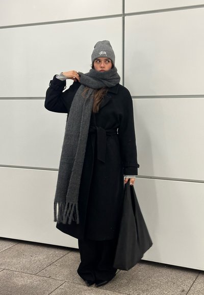 Joven mujer con abrigo y pantalones negros, gorro y bufanda de punto grises, sosteniendo un bolso tote negro, de pie contra una pared gris claro.