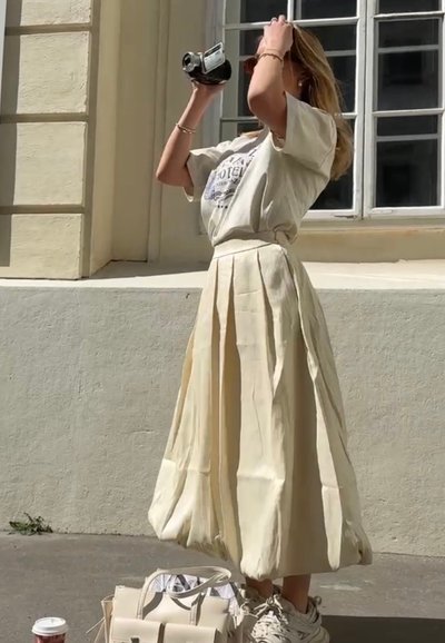 Camiseta beige de corte holgado con estampado gráfico, combinada con una falda de crema de largo hasta el suelo y con plisados. Zapatillas blancas y un bolso pequeño en la base.