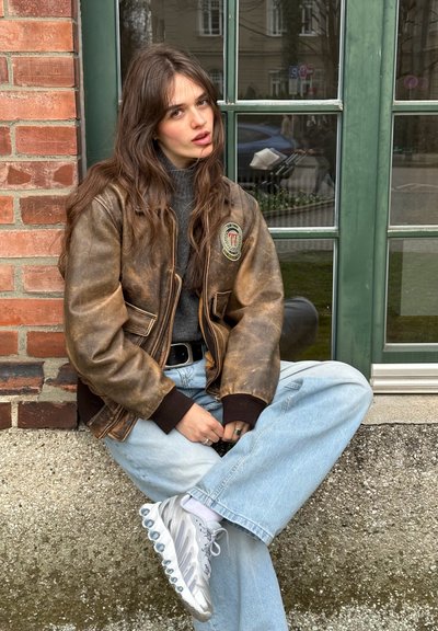 Cazadora bomber de cuero marrón con un parche, suéter gris, vaqueros baggy en azul claro y zapatillas plateadas con suelas de burbujas distintivas.