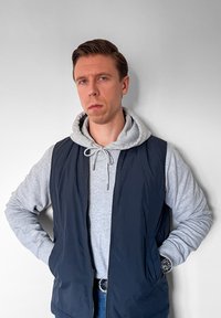 Námorně modrá zateplená vesta přes světle šedou mikinu s kapucí. Slim fit, dvoubarevný design se zipem a šňůrkou. Na zápěstí je vidět hodinky.