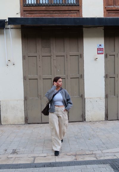 Mujer con cárdigan gris, top corto blanco y pantalones beige caminando por la acera de la ciudad con puertas cerradas marrones al fondo.